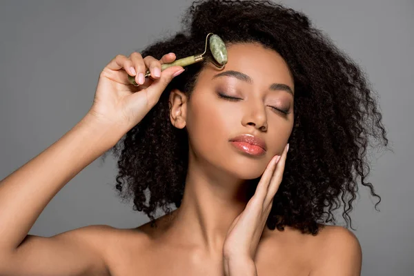 Belle fille afro-américaine avec les yeux fermés à l'aide de rouleau de jade, isolé sur gris — Photo de stock