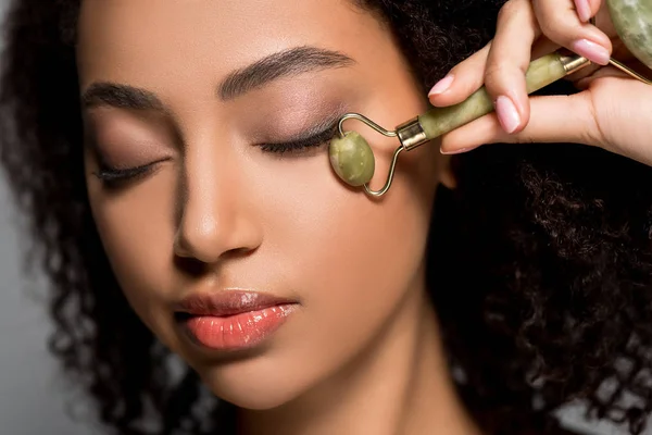 African american woman with closed eyes using jade roller, on grey — Stock Photo