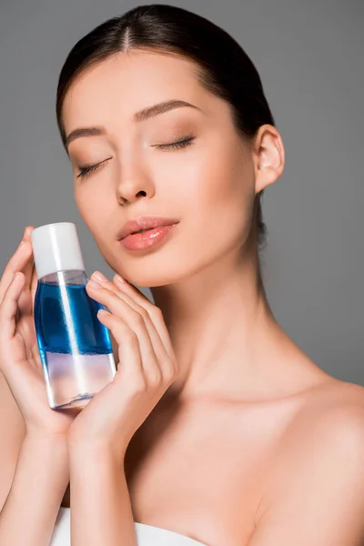 Portrait of tender woman with closed eyes holding bottle of makeup remover, isolated on grey — Stock Photo
