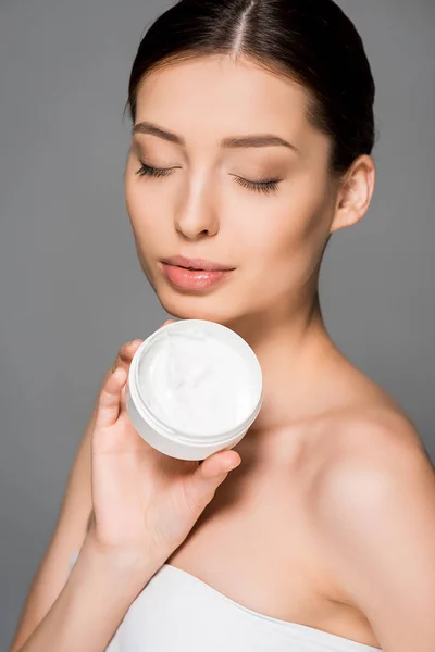 Hermosa chica con los ojos cerrados celebración crema hidratante, aislado en gris - foto de stock