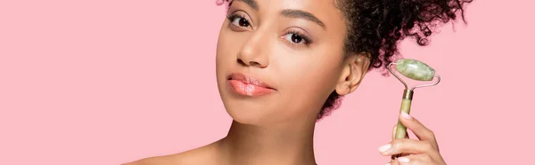 Panoramic shot of attractive african american girl using jade roller, isolated on pink — Stock Photo