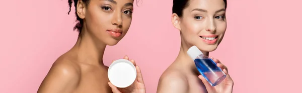 Plano panorámico de chicas multiétnicas atractivas sosteniendo crema cosmética y removedor de maquillaje, aislado en rosa - foto de stock