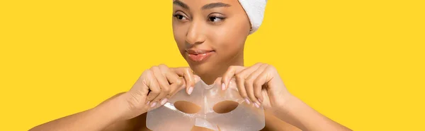 Plano panorámico de alegre chica afroamericana con toalla en la cabeza sosteniendo máscara de hidrogel, aislado en amarillo - foto de stock