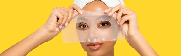 Панорамний знімок привабливої афроамериканської дівчини з рушником на голові, що застосовує гідрогелеву маску, ізольовано на жовтому — стокове фото