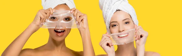 Панорамний знімок веселих мультикультурних дівчат, що тримають гідрогелеві маски, ізольовані на жовтому — стокове фото