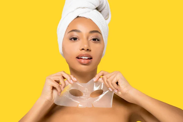 Menina afro-americana com toalha na cabeça segurando máscara hidrogel, isolado em amarelo — Fotografia de Stock