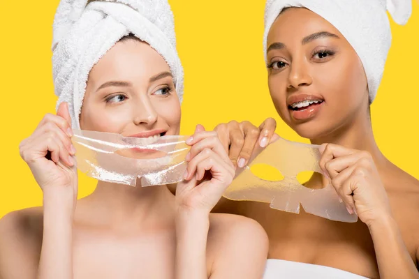 Portrait of multicultural women with towels on heads holding hydrogel masks, isolated on yellow — Stock Photo