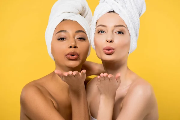 Chicas multiétnicas positivas con toallas en la cabeza soplando besos de aire, aislado en amarillo - foto de stock
