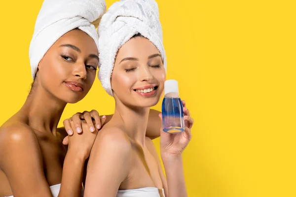 Meninas multiétnicas felizes segurando garrafa com removedor de maquiagem, isolado no amarelo — Fotografia de Stock