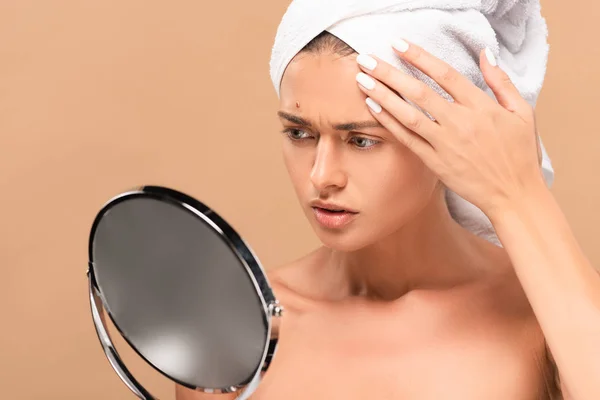 Naked woman in towel looking at mirror and touching face with pimple isolated on beige — Stock Photo