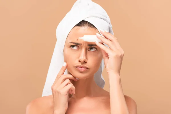 Mulher descontente e nu na toalha segurando creme de tratamento perto espinha no rosto isolado no bege — Fotografia de Stock