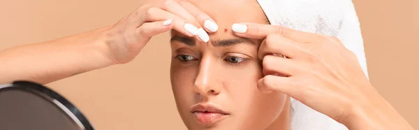 Panoramic shot of young woman in towel squeezing pimple and looking at mirror isolated on beige — Stock Photo