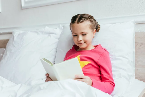 Adorable smiling child reading book while sitting on bed — Stock Photo
