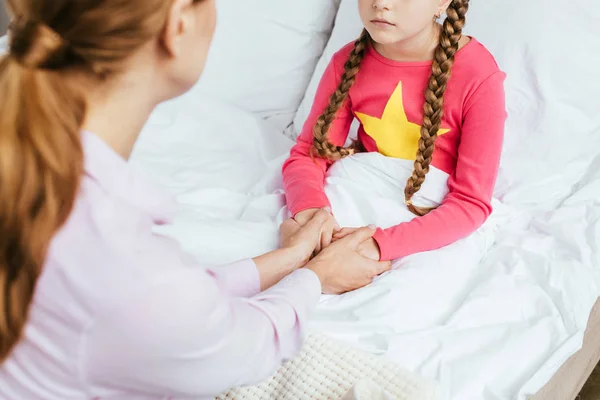 Vista recortada de la madre cogida de la mano con hija enferma en la cama - foto de stock