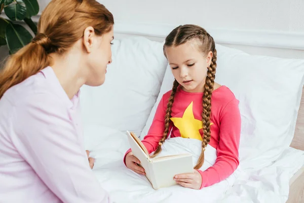 Mignonne fille lecture livre à mère dans le lit — Photo de stock