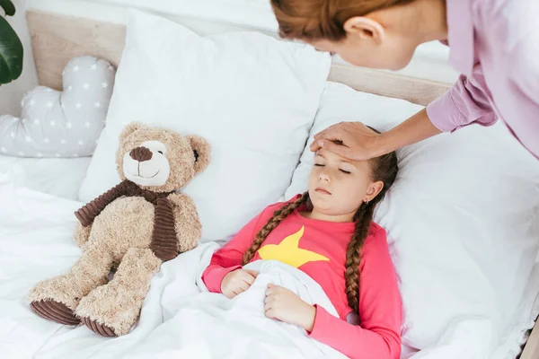 Mother touching forehead of ill sleepy daughter in bed with teddy bear — Stock Photo