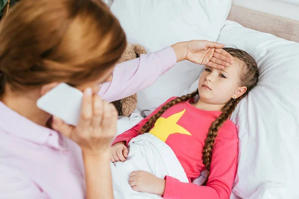 Worried mother talking on smartphone while touching forehead of ill daughter with fever — Stock Photo