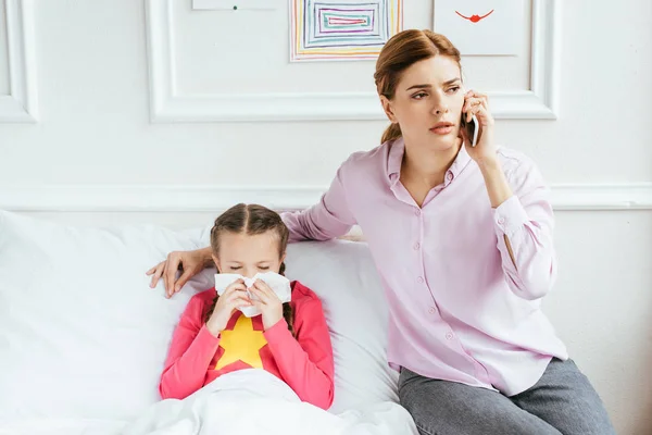 Worried mother having online consultation on smartphone while sick daughter with runny nose sitting in bed — Stock Photo
