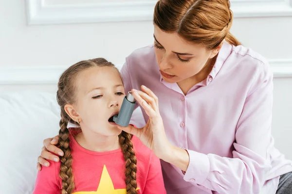 Hija triste con asma usando inhalador con mamá preocupada cerca - foto de stock