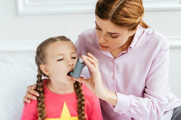 Sad daughter with asthma using inhaler with worried mom near — Stock Photo