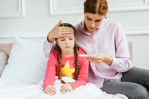 Mutter blickt auf Thermometer, während sie Stirn kranker Tochter mit Fieber berührt — Stockfoto