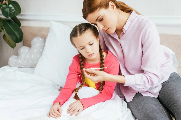 Madre mirando el termómetro con hija enferma en la cama - foto de stock