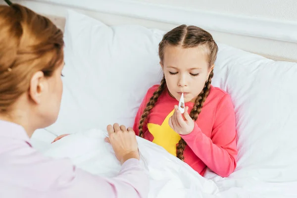 Erkrankte Tochter misst Fieber mit Thermometer, während sie mit Mutter im Bett sitzt — Stockfoto