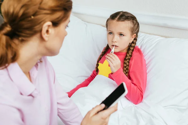 Hija enferma midiendo la fiebre con termómetro mientras la madre tiene consulta en línea en el teléfono inteligente — Stock Photo