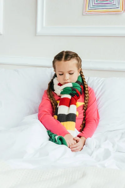 Triste niño enfermo en bufanda sentado en la cama - foto de stock