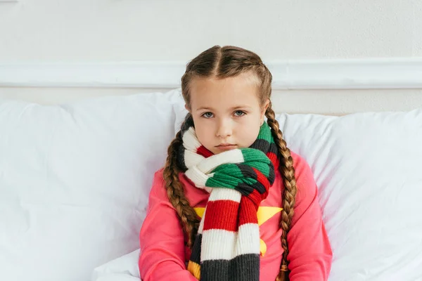 Грустный больной ребенок в шарфе сидит на кровати — стоковое фото