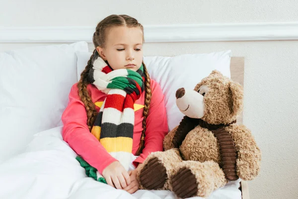 Triste criança doente no cachecol sentado na cama com ursinho de pelúcia — Fotografia de Stock