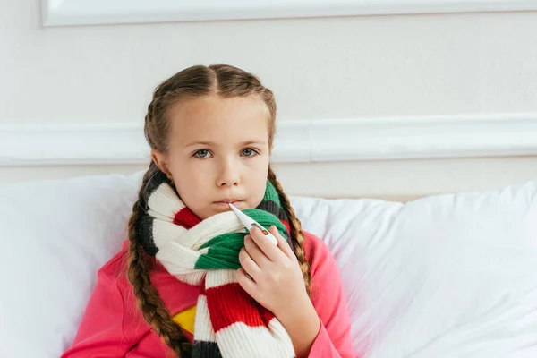 Sad ill kid in scarf having fever and holding thermometer on bed — Stock Photo