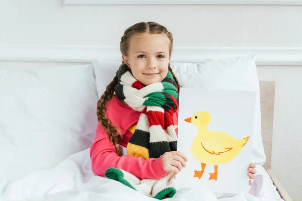 Smiling sick kid in scarf holding painting with duck on bed — Stock Photo