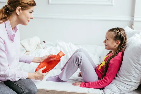 Smiling mother giving hot water bottle to ill daughter sitting on bed — Stock Photo