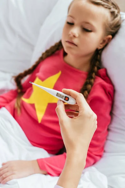 Cropped view of mother looking at thermometer with sick daughter in bed — Stock Photo