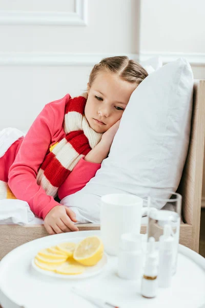 Грустный больной ребенок в шарфе лежит на кровати с лекарствами рядом — стоковое фото