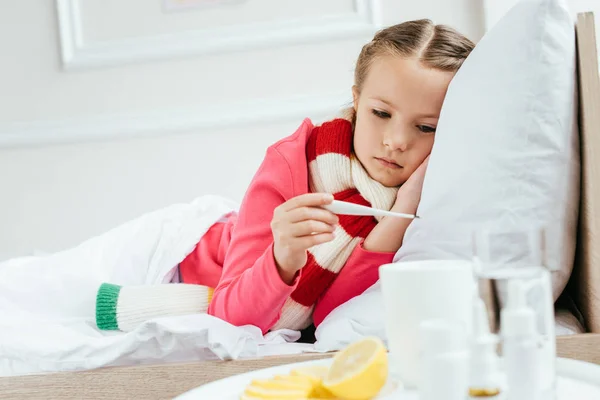 Больной расстроенный ребенок с лихорадкой в шарфе глядя на термометр во время лежания на кровати с лекарствами рядом — стоковое фото