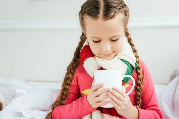 Cute ill kid in scarf holding cup of hot drink while sitting on bed — Stock Photo