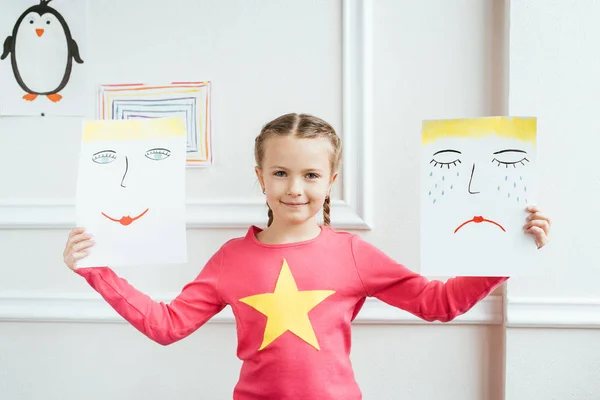 Niño alegre sosteniendo dos cuadros con caras felices y tristes - foto de stock