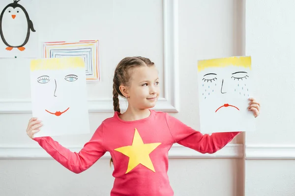 Улыбающийся ребенок, держащий две картины с счастливыми и грустными лицами — стоковое фото