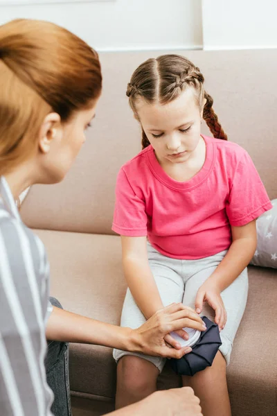 Mom holding ice bag compress on painful knee of sad daughter — Stock Photo