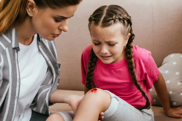 Mère tenant la jambe avec blessure au genou tandis que sa fille pleure à la maison — Photo de stock