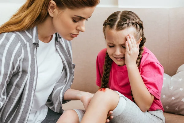 Madre sosteniendo la pierna con herida en la rodilla mientras hija emocional llorando en casa - foto de stock