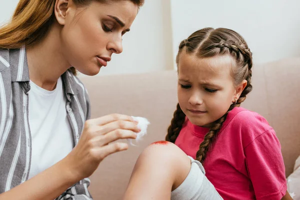 Mother holding antiseptic cotton near wound on knee of sad daughter — Stock Photo