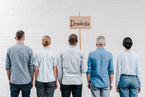 Vue arrière des personnes multiculturelles debout près du mur de briques et de la plaque avec lettrage demokratie — Photo de stock