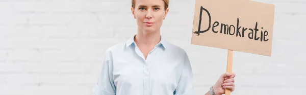 Panoramic shot of woman holding placard with demokratie lettering near brick wall — Stock Photo