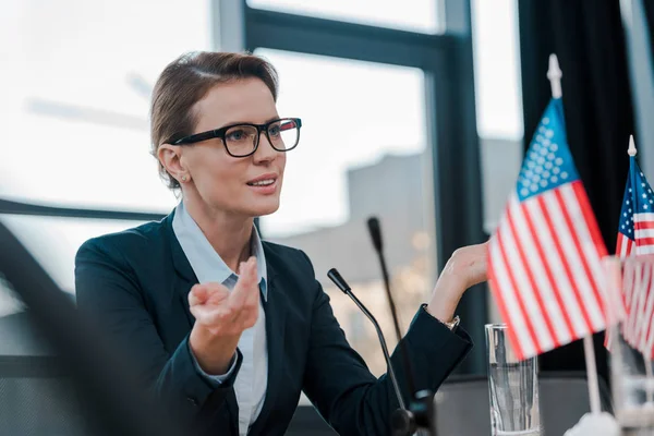 Beautiful diplomat in eyeglasses gesturing while talking near microphone and american flags — Stock Photo