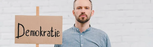 Panoramic shot of bearded citizen holding placard with demokratie lettering near brick wall — Stock Photo