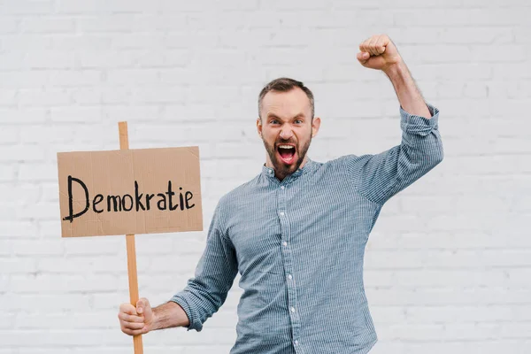 Emotional citizen holding placard with demokratie lettering and screaming near brick wall — Stock Photo