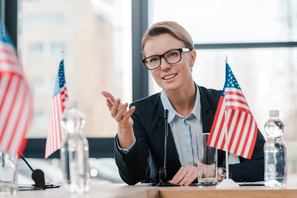 Smiling diplomat in eyeglasses gesturing while talking near microphone and american flag — Stock Photo